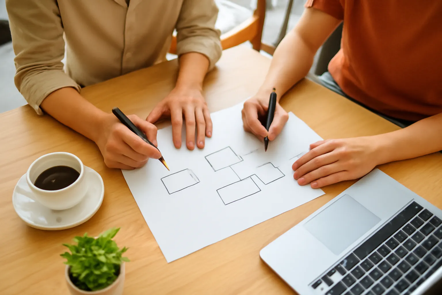 Two colleagues sketching boxes and arrows on blank paper at a wooden cafe table